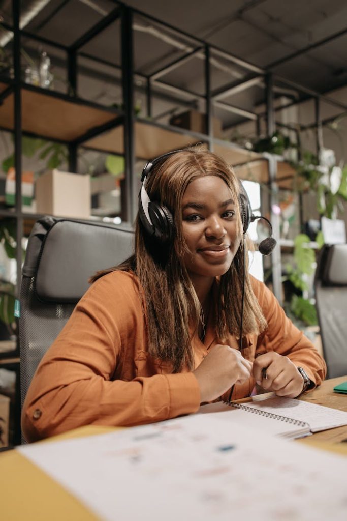 VA Services - a-woman-with-headphones-sitting-at-a-desk-with-a-notebook-7658382 VA Services - Smiling call center agent with headphones working indoors, sitting at a desk.